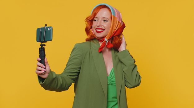 Redhead woman in jacket traveler blogger taking selfie on mobile phone selfie stick, communicating video call online with subscribers. Young ginger girl isolated alone on yellow studio background