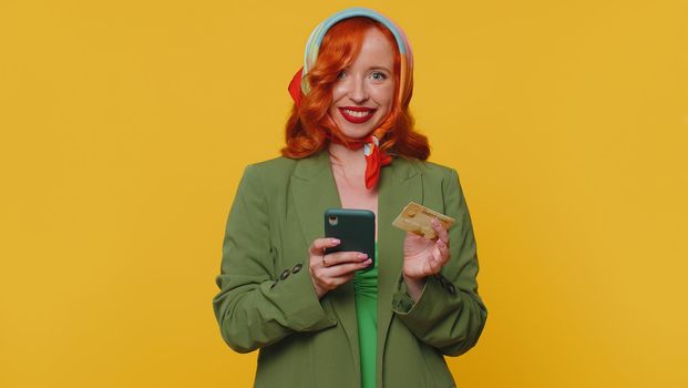 Sincere redhead woman customer using credit bank card and smartphone while transferring money, purchases online shopping, payment. Internet finance. Young girl isolated alone on yellow wall background