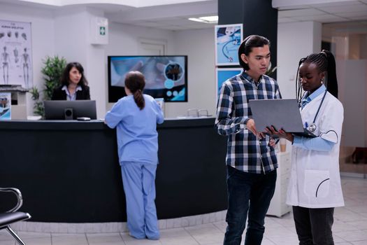African american medical doctor holding laptop presenting lab results to asian patient in private practice clinic. Medic talking with young man checking appointment on portable computer.