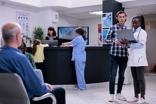 African american doctor holding laptop with mri scan results talking with asian patient in busy private clinic waiting room. Medic having conversation with young man checking appointment.