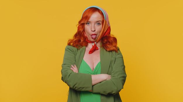 Funny joyful sincere redhead woman 20s in jacket making playful silly facial expressions and grimacing, fooling around showing tongue. Young beautiful ginger girl isolated alone on yellow background