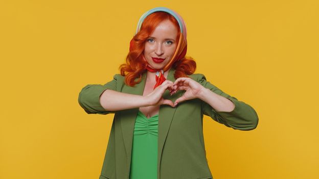 Girl in love. Smiling redhead woman in green dress makes heart gesture demonstrates love sign expresses good feelings and sympathy. Young one ginger girl isolated alone on yellow studio background