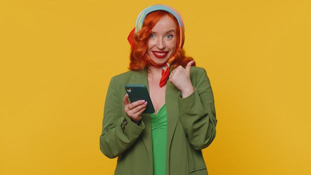 Beautiful red hair woman using mobile phone typing new post on web sms message, browsing, searching addiction of social networks. Young ginger girl studio shot isolated alone on yellow wall background