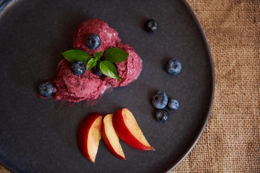 Flat lay of three purple frozen balls of organic blueberry sorbet, decorated with blueberries, lemon basil leaf and slices of ripe juicy peach. Healthy Live Food. Veganism Raw Food. Dieting concept