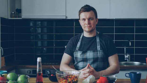 Portrait of young handsome caucasian executive chef in apron sitting at table in modern lighty spacious kitchen in front of plates , he is smiling, calm and positive, looking at camera