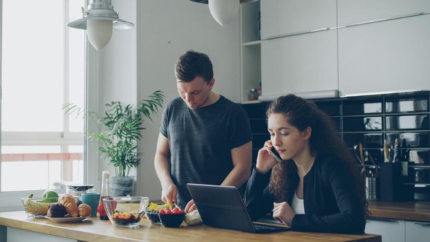 serious caucasian nice couple in kitchen, man is cutting salad, he is standing silent, woman is sitting at table working on laptop computer and talking on phone