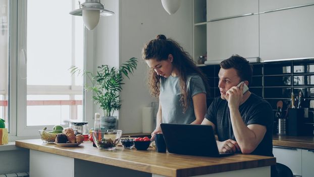 serious couple in kitchen, woman is cutting salad, she is standing silent, man is sitting at table working on laptop and talking on phone, he is annoyed and angry