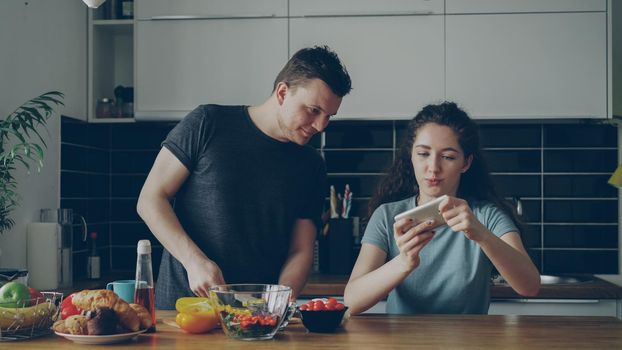 Attractive couple in the kitchen at home. Curly girl playing video game on smartphone while her boylfriend cooking breakfast