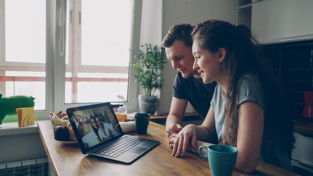 Young caucasian couple sitting at table in front of laptop talking with their happy positive tanned friends from abroad