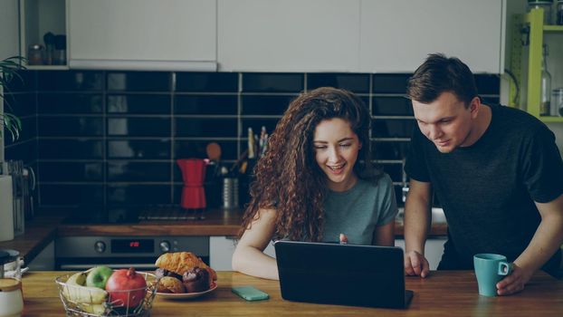 Young positive caucasian couple sitting at table in kitchen using laptop smiling and discussing at home