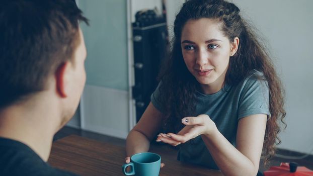 young caucasian couple sitting at table in modern spacious, kitchen dicussing something in positive way, man is sitting back to camera they are holding cups drinking tea, pretty woman is smiling