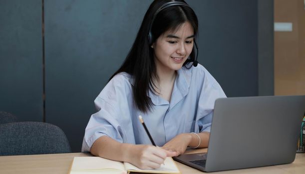 Smiling Asian young female using headset looking at laptop screen listen and learning online courses. Happy chinese business woman with headphones video call for customer service.