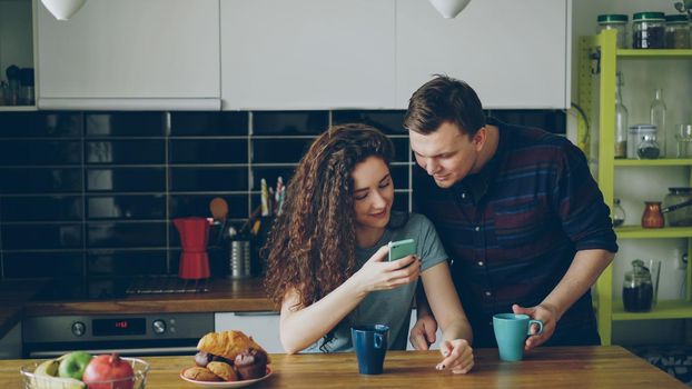 young attractive curly happy caucasian woman is sitting in modern kitchen indoors texting someone via smartphone, her husband comes and she shows something funny in her phone
