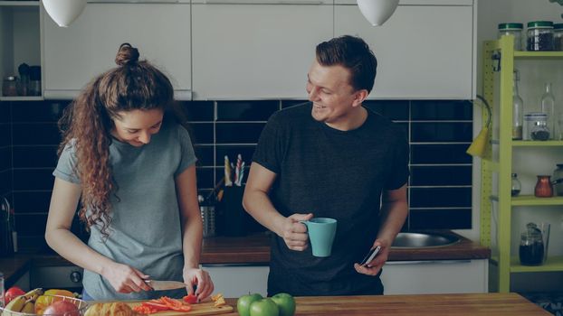 Curly woman cooking near table cutting red pepper in kitchen at home, her boyfriend showing her something funny in smartphone and they are smiling