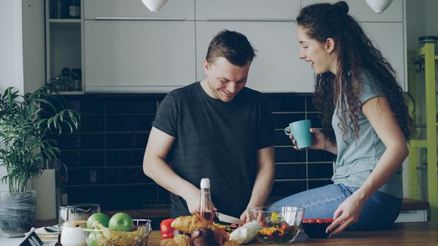 Happy young couple cooking and chatting happily while man cutting vegetables for healthy salad in the kitchen at home. Relationship and family concept