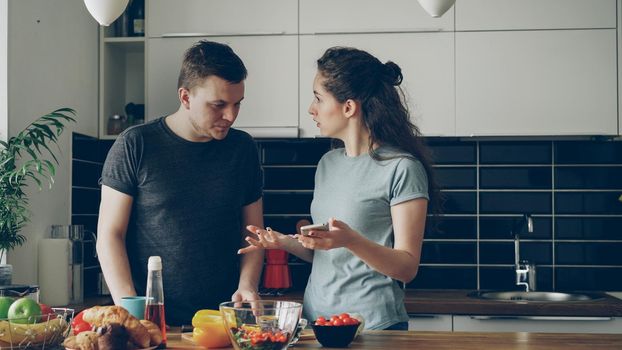 young curly caucasian woman showing something unpleasant in husband's phone while he is cooking , they are shouting and quarrelling, man is angry and irritated
