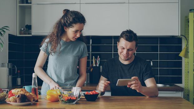 Attractive couple in the kitchen. Man playing video game on smartphone while his girlfriend cooking breakfast early morning at home