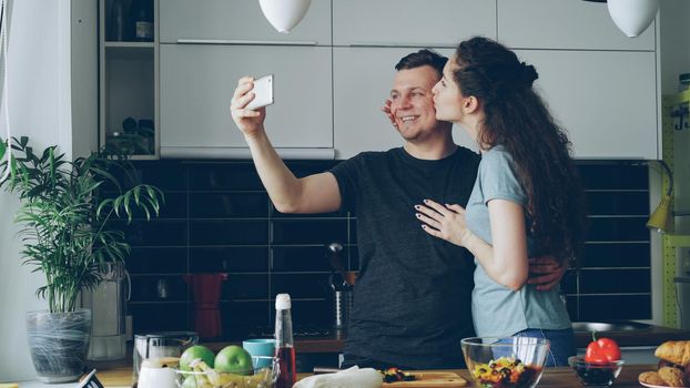 Young funny couple taking self portrait with smartphone camera while cooking in the kitchen at home in the morning