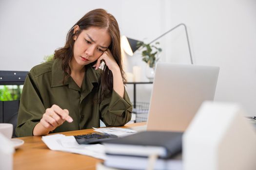 Stressed over bills. Asian young woman with laptop holding papers in her hands she calculating dealing financial budget on desk, female worried have stress headache after calculate cost, money problem