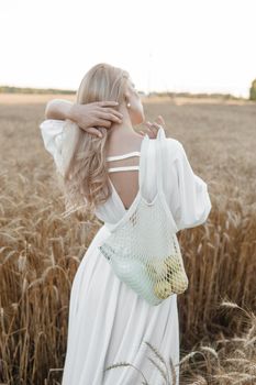 A blonde woman in a long white dress walks in a wheat field. The concept of a wedding and walking in nature.