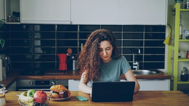young pretty curly serious caucasian woman sitting alone at table in nice spacious modern kitchen idoors working on laptop