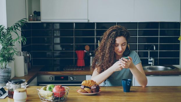 young beautiful curly pretty caucasian woman sitting at table in nice modern kitchen using smartphone, she is texting someone and smiling, calm, happy and positive