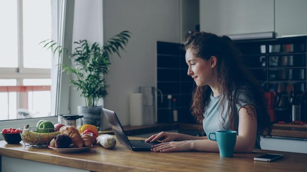 Young concentrated cheerful woman working in kitchen typing laptop computer during breakfast in the morning at home
