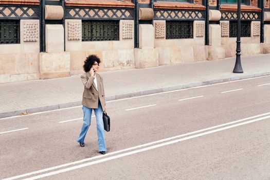 latin business woman walking across the street talking by phone with a briefcase in her hand, concept of urban lifestyle and success, copy space for text