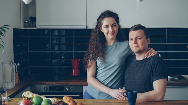 portrait of young positive smiling caucasian couple in kitchen, beautiful woman is standing near handsome man, she is hugging him, they are looking into camera