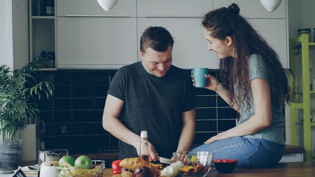 Happy young couple cooking and chatting happily while man cutting vegetables for healthy salad in the kitchen at home. Relationship and family concept
