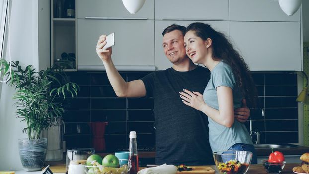 young happy couple having online video call with smartphone camera while cooking in the kitchen at home