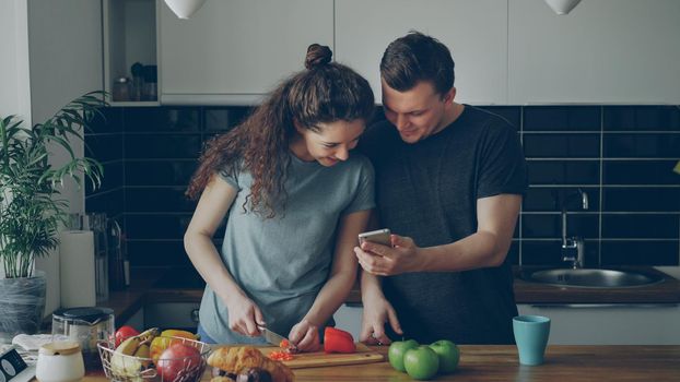 Curly woman cooking near table cutting red pepper in kitchen at home, her boyfriend showing her something funny in smartphone and they are smiling