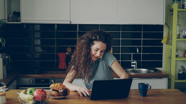 Young curly pretty positive attractive caucasian woman working on laptop and talking on phone sitting in modern light kitchen, she is printing and talking the same time