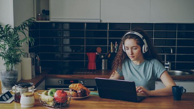 positive happy caucasian pretty curly girl sitting at table chatting with someone on laptop listening music and dancing in headphones in modern kitchen indoors