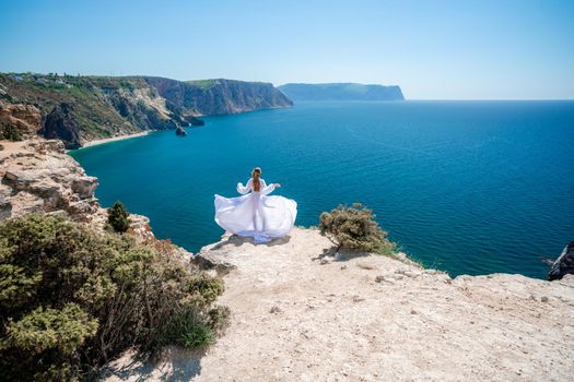 Blonde with long hair on a sunny seashore in a white flowing dress, rear view, silk fabric waving in the wind. Against the backdrop of the blue sky and mountains on the seashore