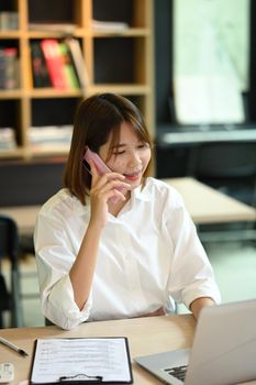 Charming working woman having phone conversation with her business partner while sitting in modern workplace.
