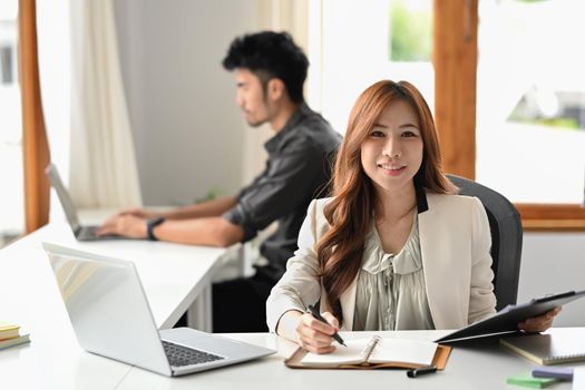 Attractive asian woman employee sitting front of laptop at corporate office and smiling to camera.