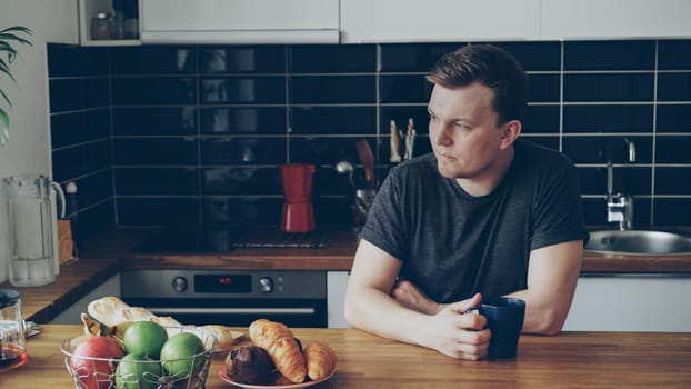 Portrait of young caucasian handsome man sitting at table in modern spacious kitchen, looking through window, drinking tea, he is tired and upset