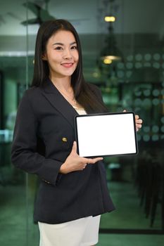 Confident millennial asian woman in business suit holding digital tablet with empty screen and smiling to camera.