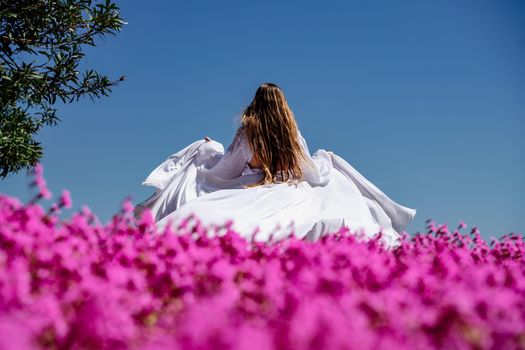 a beautiful woman in a white flowing long dress stay near a beautiful field with pink flowers