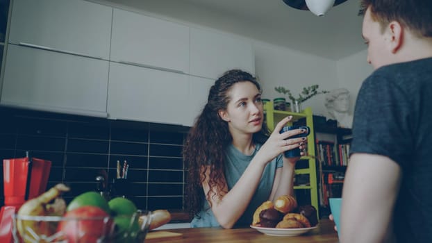 young caucasian couple sitting at table in modern kitchen dicussing something, curly woman is sitting back to camera drinking tea and eating croussant, they are holding cups