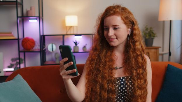 Smiling young redhead woman wear earphones making smartphone conversation call with friends sitting at home. Happy excited ginger businesswoman girl enjoying mobile phone loudspeaker talking indoors
