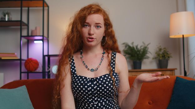 POV of redhead young woman sitting on couch, looking at camera, making video webcam conference call with friends or family, enjoying pleasant conversation. Girl laughing, waving hello alone in home
