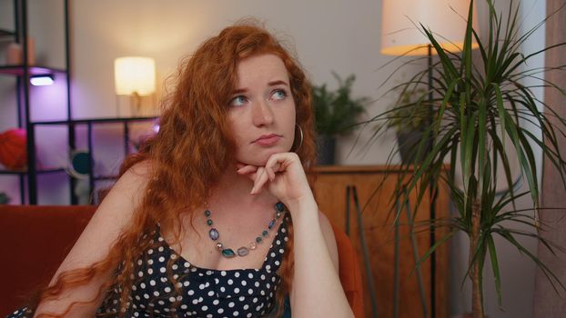 Close-up of sad young redhead woman sitting at home looks pensive thinks over life concerns or unrequited love, suffers from unfair situation. Problem, break up, depressed feeling bad annoyed, burnout