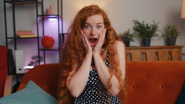 Oh my God, Wow. Redhead young woman surprised looking at camera with big eyes, shocked by sudden victory, good win news, celebrating at home. Excited amazed ginger girl indoors in living room on couch