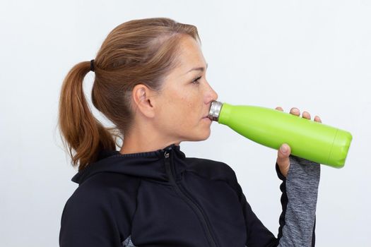 Caucasian middle aged woman in sportiv dress holding light green thermos bottle drinking water on white background