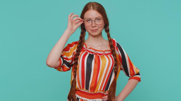 Cheerful lovely redhead girl in colorful dress smiling, looking at camera. Young fashion model teenager child kid wearing glasses indoor studio shot isolated on blue background. Female nature beauty