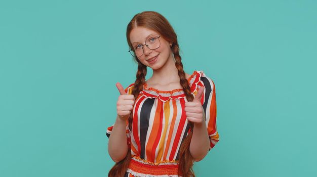 Redhead young teen girl in dress raises thumbs up agrees with something or gives positive reply recommends advertisement likes good. Ginger teenager child kid isolated alone on blue studio background