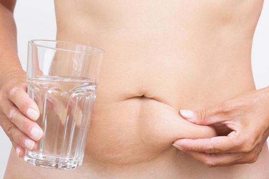 Cropped woman body with hand touching fat on belly and hand holding glass of pure water on white background. Concept water and weight loss
