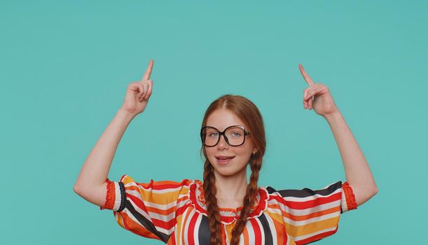Beautiful redhead girl kid showing thumbs up and pointing up empty place, advertising area for commercial text, copy space for goods promotion. Young ginger teenager child on blue studio background